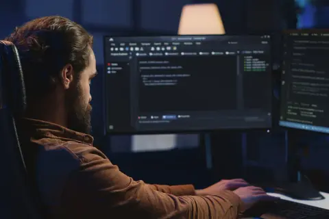 Mężczyzna z brodą, jako java developer, intensywnie pracuje przy dwóch monitorach, pisząc kod w ciemnym pomieszczeniu.