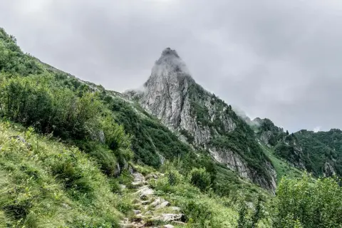 Sentier rocailleux grimpant vers un pic montagneux enveloppé de brume. Végétation luxuriante sur les pentes.