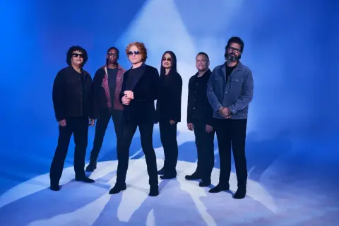 Zespół Simply Red gotowy na koncert. Muzycy pozują na tle niebieskiego światła, tworząc atmosferę nadchodzącego wydarzenia.