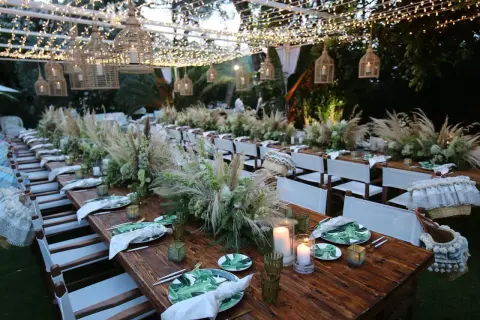 Tavoli in legno apparecchiati per un matrimonio in campagna, con centrotavola di fiori e pampas, luci soffuse e cesti decorativi.
