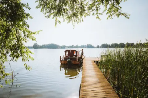 Ein Mann steht auf einem Holzsteg neben einem Hausboot auf einem See. Perfekter Urlaub in Brandenburg.