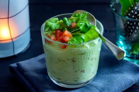 Krämig guacamole med crème fraîche, toppad med färsk koriander och tärnad tomat. Serveras i ett glas med en sked.
