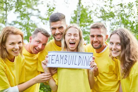 En grupp glada människor i gula tröjor håller upp en skylt med "TEAMBUILDING". De verkar ha det roligt, som om de precis svarat på några snabba roliga frågor.