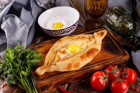 Chaczapuri adżarskie z jajkiem i serem, podane na desce z pomidorami i natką pietruszki. Idealne na kazdy posiłek.