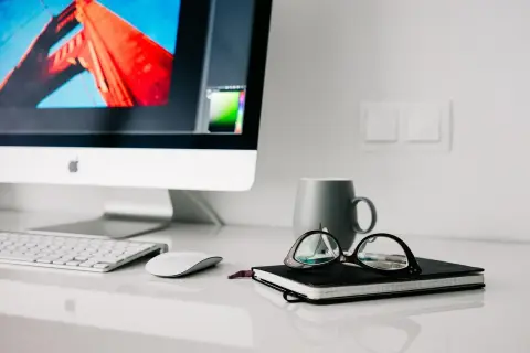 Na biurku iMac, klawiatura, myszka, notatnik z okularami i kubek. Higiena pracy to dbanie o porządek i komfort.