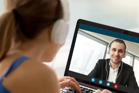 Kobieta w słuchawkach pisze na laptopie, prowadząc wideokonferencję z uśmiechniętym mężczyzną. On też ma słuchawki, ale ona pisze ale się nie umawia.