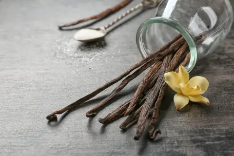 Un tas de gousses de vanille séchées débordent d'un bocal en verre, accompagnées d'une fleur de vanille jaune et d'une cuillère de sucre.