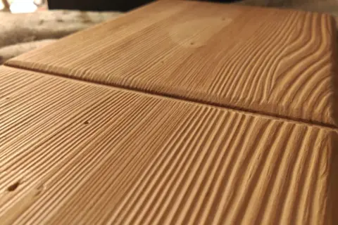 Superficie de madera con surcos paralelos que resaltan las vetas naturales. La luz crea sombras que dan profundidad a la textura.