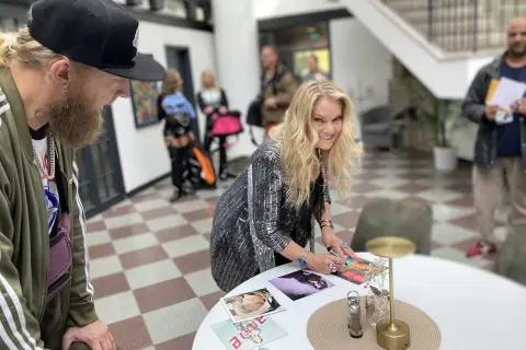 Ankie Bagger signerar bilder vid ett bord. En man med skägg och keps står bredvid.