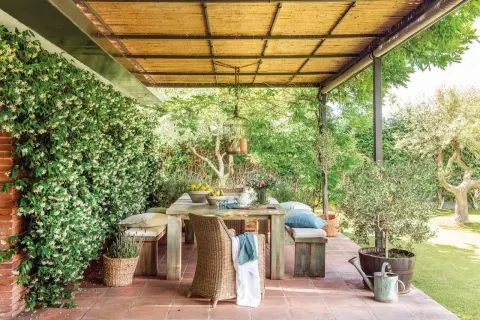 Un porche de cañizo con una mesa de madera rústica, sillas de mimbre y bancos, rodeado de vegetación exuberante y flores blancas.