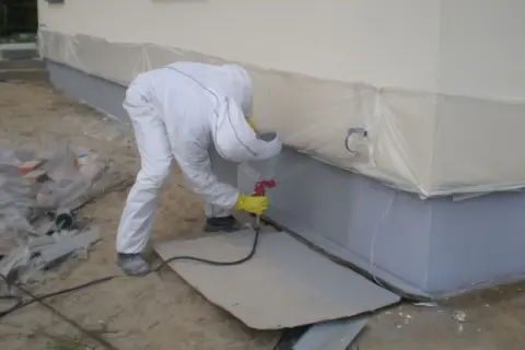 Pracownik w kombinezonie ochronnym wykonuje malowanie elewacji, natryskując szary tynk na cokół budynku.