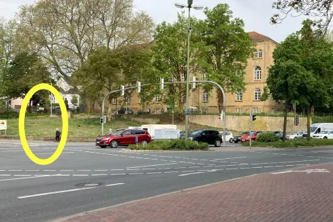 Eine Person wartet an der Ampel, um links abzubiegen. Autos stehen bereit, während im Hintergrund ein Gebäude und Bäume zu sehen sind.