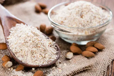 Cuillère en bois remplie de poudre d'amande, idéale pour remplacer la farine dans vos recettes. Amandes entières et bol de poudre à côté.