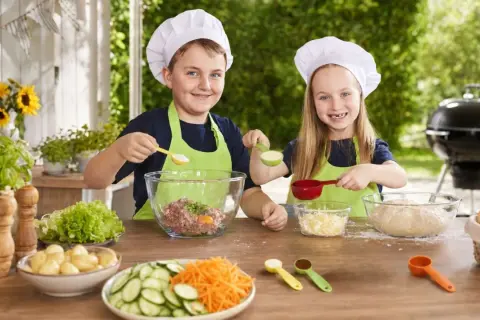 Zwei Kinder mit Kochmützen bereiten sich auf das Grillen vor. Sie mischen Zutaten für Burger und haben Spaß beim gemeinsamen Kochen.