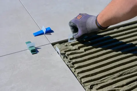 Pose de carreaux : un gant ajuste un coin pour enlever colle carrelage sèche. Des cales vertes et bleues sont déjà en place.
