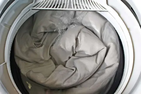 A grey comforter is being washed in a washing machine, showing the best way to wash a comforter. Water is splashing onto the fabric.