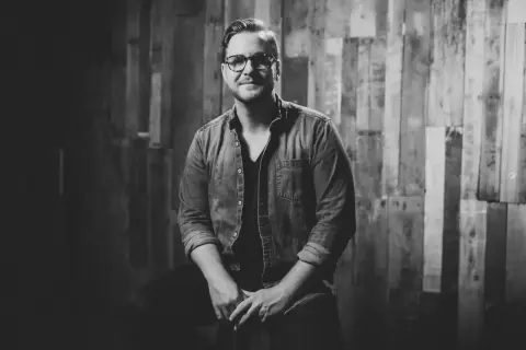 Jordan Reynolds, a talented songwriter, poses confidently in a denim shirt against a rustic wooden backdrop.