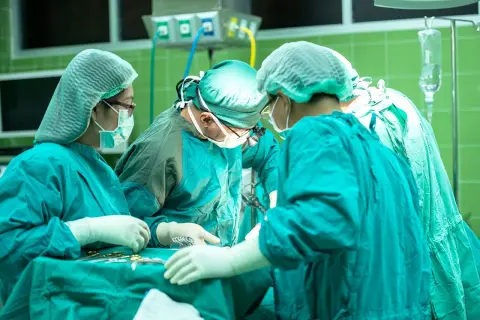 Zespół chirurgów w sterylnych strojach pracuje przy stole operacyjnym. W tle widać aparaturę medyczną. Sytuacja budzi niepokój, czy nieumyślne spowodowanie śmierci nie jest tu możliwe.