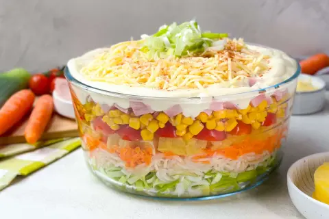 Omas Schichtsalat in einer Glasbowle, geschichtet mit Lauch, Karotten, Ananas, Tomaten, Mais und Schinken, gekrönt mit Mayonnaise, Käse und Frühlingszwiebeln.