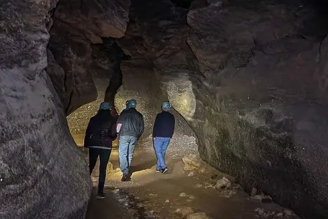 Tres personas con cascos exploran una cueva oscura, sus linternas iluminan las paredes rocosas y el suelo irregular. Los yesos naturales brillan en la oscuridad.