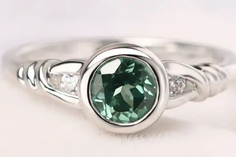 Anillo de plata con piedra verde ovalada y dos pequeños diamantes a cada lado, sujeto con un engaste bisel.