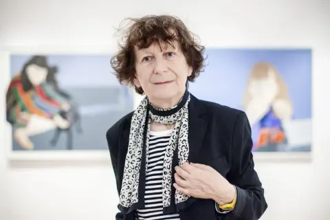Ewa Kuryluk, artystka o burzy brązowych włosów, w czarnej marynarce i pasiastym szalu, pozuje na tle swoich obrazów.