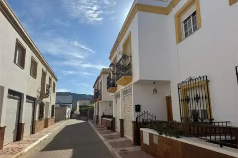 Calle soleada con casas blancas y detalles amarillos en Celin, Almería. Balcones de hierro forjado y un cielo azul con nubes.