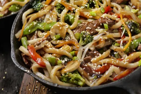 Spaghetti di soia con verdure miste, carne e semi di sesamo in una padella scura.