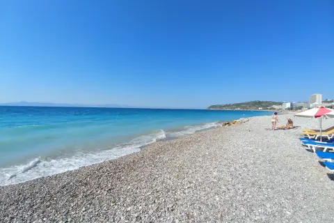 Słoneczna plaża w Ixia, Rodos, z turkusowym morzem i kamienistym brzegiem. Dwie osoby odpoczywają pod parasolem.