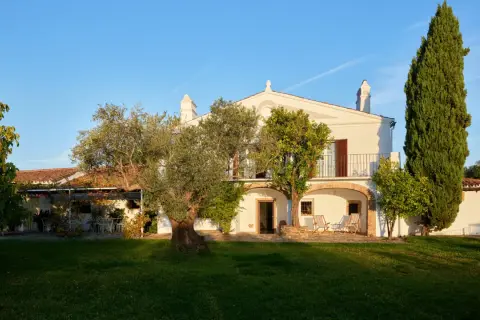 La finca el azahar, una casa blanca con balcón y arcos, rodeada de olivos y cipreses bajo un cielo azul.