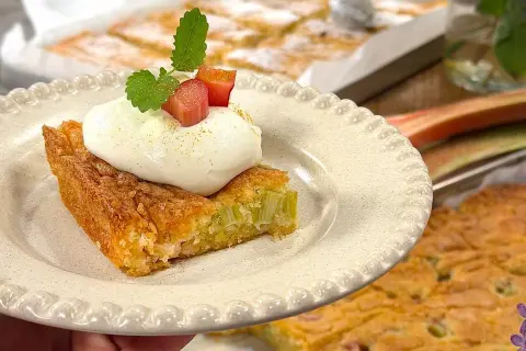 En bit saftig rabarberkaka i långpanna, toppad med fluffig grädde och färska rabarberbitar. Perfekt till fikat!