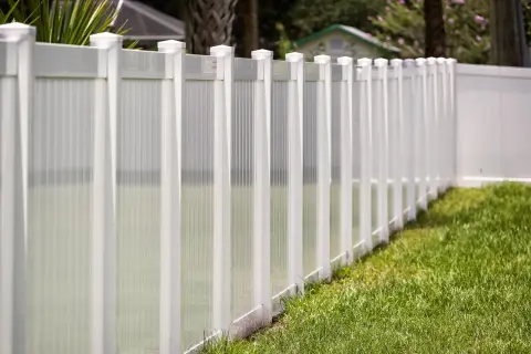 A newly completed vinyl fence installation, featuring white posts and translucent panels, lines a lush green lawn.