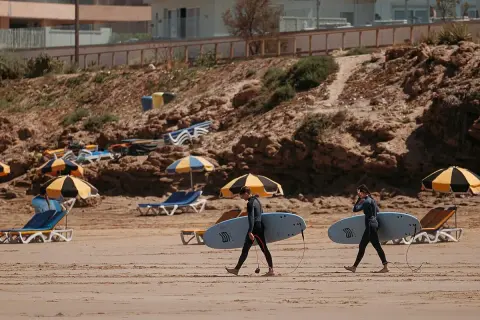 Deux surfeurs marchent sur la plage de Tamraght, Maroc, avec leurs planches sous le bras. Des parasols et des chaises longues sont installés sur le sable.