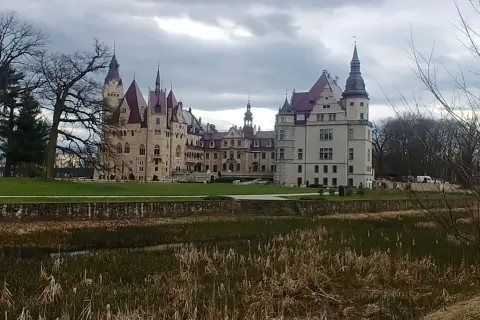 Majestatyczny zamek w Głogówku z wieżami i czerwonymi dachami, otoczony zielonym trawnikiem i stawem z trzcinami.