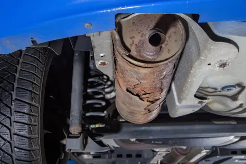 Vue du dessous d'une voiture bleue, montrant une roue, un ressort de suspension et un silencieux rouillé et endommagé.