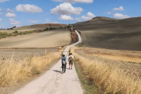 Due pellegrini camminano lungo la Via Francigena, immersi in un paesaggio collinare toscano sotto un cielo azzurro con nuvole bianche.