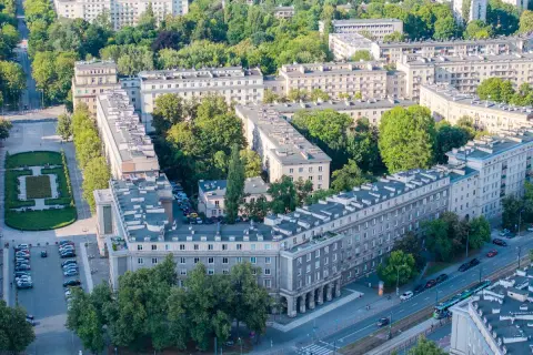 Osiedle w stylu socrealistycznym, przypominające architekturę Nowej Huty, z zielenią i ulicą z tramwajem.