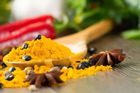 Une cuillère en bois déborde de poudre de curcuma, parsemée de grains de poivre noir et d'anis étoilé. Un **tabel karouia** pour une cuisine pleine de saveurs.