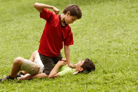 Un niño en camiseta roja agrede a otro en el suelo. Un ejemplo de cómo los niños que excluyen a otros pueden causar daño.