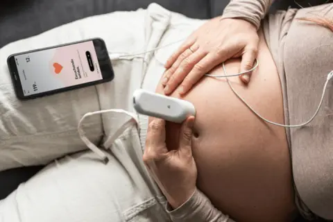 Manos de mujer embarazada usando un doppler fetal para escuchar el latido del bebé. La pantalla del móvil muestra "Escuchando latidos".