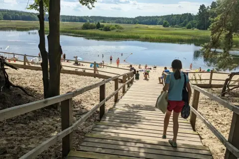 Dziewczynka schodzi drewnianymi schodami na piaszczystą plażę nad jeziorem. W oddali widać ludzi kąpiących się i odpoczywających. Stawy echo zwierzyniec to idealne miejsce na letni relaks.