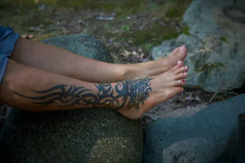 Beine mit einem Tribal-Tattoo, das sich über Wade und Fuß zieht. Das Tattoo kribbelt, als die Person auf einem Stein sitzt.