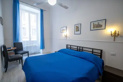 Pokój z niebieską pościelą, idealny na tanie noclegi w Rzymie u Polaków. Okno z niebieskimi zasłonami, dwa krzesła i stolik.