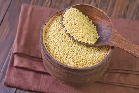 Miseczka pełna kaszy jaglanej, która nie zawiera glutenu. Drewniana łyżka z ziarnami na brązowej serwetce.