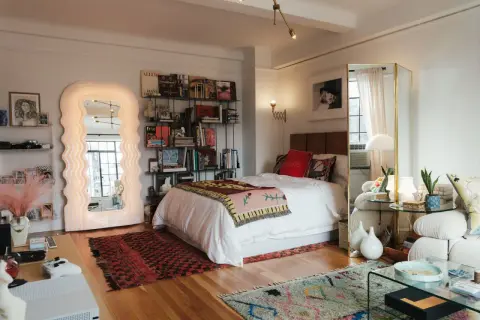Un dormitorio con un diseño estudio moderno. Cama blanca, estanterías llenas de libros, un espejo ondulado y alfombras coloridas.