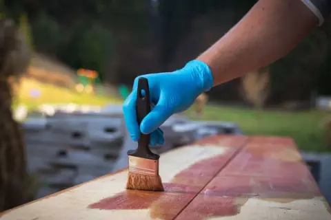 Main tenant une brosse, applique une lasure sur des planches de bois à l'extérieur.