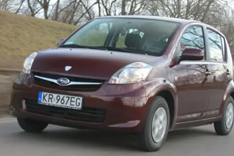Ciemnoczerwony Subaru Justy jedzie po drodze, zalesione wzgórze w tle.