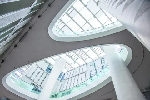 Nowoczesne wnętrze z białymi kolumnami i szklanymi balustradami, przez które wpada obfite światło dzienne.