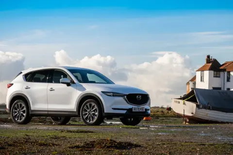 Vit Mazda CX-5 parkerad vid en båt på en lerig strand, med hus och moln i bakgrunden.