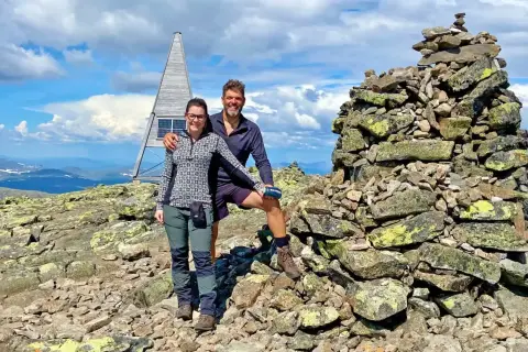 Två glada vandrare vid en stenröse på en bergstopp i Sverige. En modern, pyramidformad byggnad med solpaneler syns i bakgrunden. Perfekt för att utforska Sveriges vandringsleder.